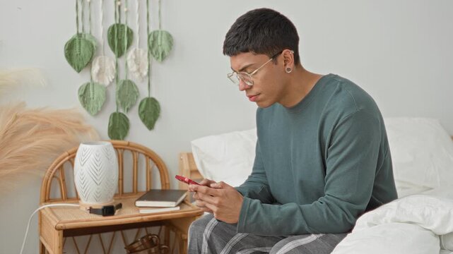 Young latino man sitting on bed holding smartphone with hands covering face in bedroom, wearing green sweater and plaid pants; anxiety solitude.