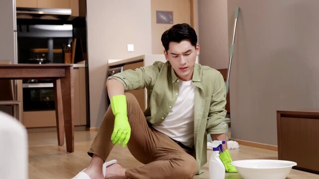 Unhappy tired man in rubber gloves sitting on the ground after doing household chores washing floor using spray detergent supplies cleaning, wrings rag. Housekeeping, household chores, maid work job