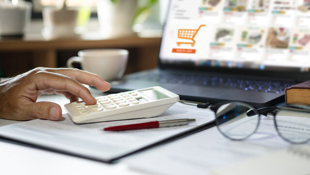 close-up shot, man hand using calculator to calculate cost, profit after start new business at home via online platform on laptop, online shopping and home shopping concept
