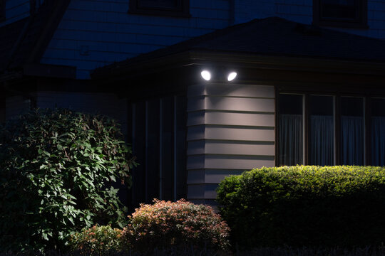 Bright LED floodlights illuminate the corner of a modern house at night, casting light on lush green and reddish bushes, creating a secure and inviting evening atmosphere.