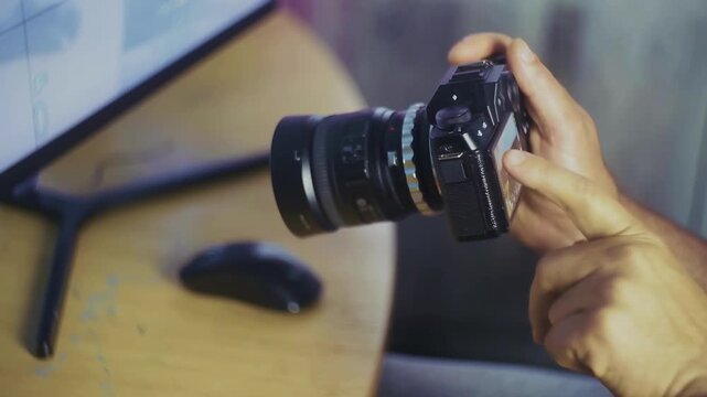 A person holds a DSLR camera with a lens, seemingly preparing to take a photo. In the background, a computer monitor and mouse sit on a wooden desk.