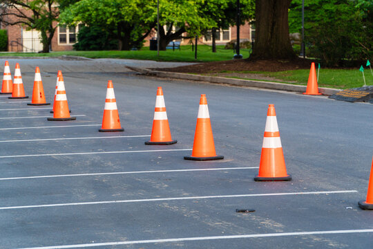 A line of bright orange traffic cones with white stripes on dark asphalt pavement, defining a temporary barrier in a parking lot, with lush green trees and buildings in the background, suggesting.