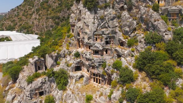 Demre, Turkey. Lycian rock-cut tombs of ancient Myra, house-tomb facades with columned porticoes and pediments carved into sheer limestone cliffs, aerial view under bright summer sun. Aerial View, Sp