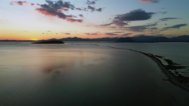 Aerial drone tracking shot following a scenic coastal road and winding pathways of Koronisia, moving towards a dramatic golden sunset over the Ambracian Gulf. High quality 4K stock footage.