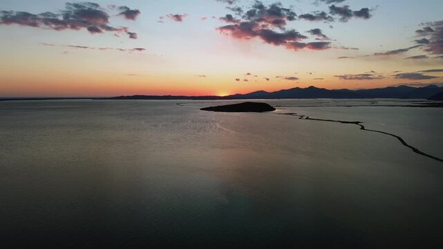Panoramic aerial shot flying over the serene, reflective waters of the Ambracian Gulf in Koronisia, Greece, facing a vibrant sunset horizon with a small island silhouette. High quality 4K video.
