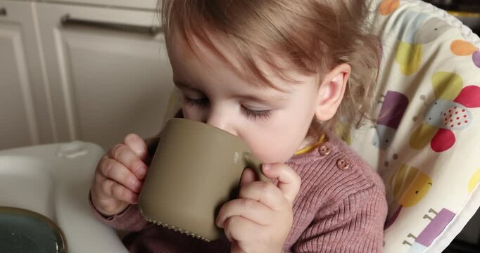 Little child sitting in highchair drinking from silicone cup with two handles. Cute toddler girl holding mug drinking water milk juice independently kitchen interior background. Care complementary