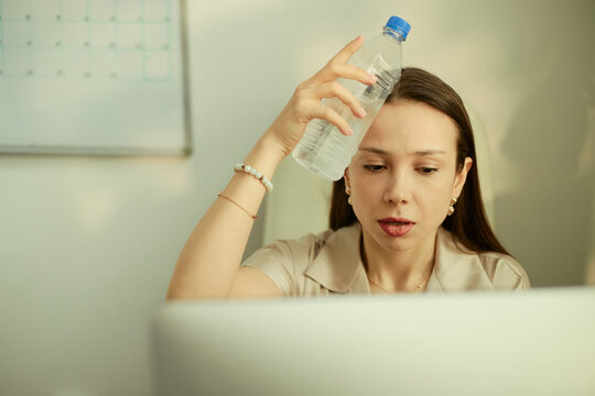 Young adult Caucasian businesswoman cooling forehead with water bottle while working on laptop, showing heat stress and fatigue. Useful for workplace wellness, summer office safety
