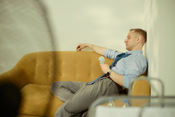 Caucasian young businessman resting on sofa and holding portable fan, showing office fatigue and...
