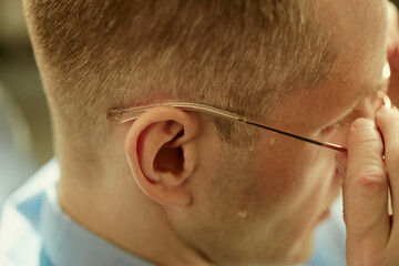 Fototapeta premium Close up of Caucasian young adult man adjusting eyeglasses and rubbing eyes, showing eye strain and fatigue. Suitable for vision care, workplace stress, burnout, or screen fatigue campaigns