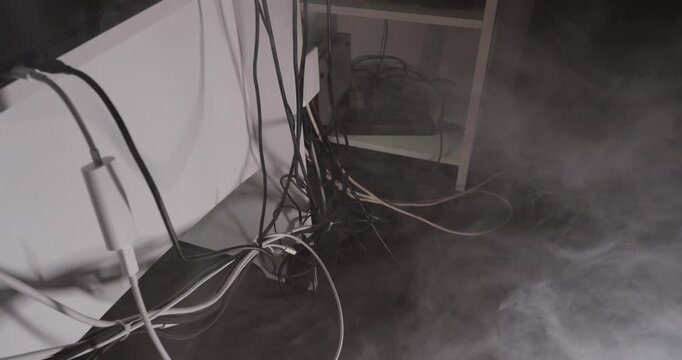 showing smoke rising from tangled electrical cables behind furniture, representing cable overheating, electrical malfunction, fire risk, and household safety hazards. Suitable for electrical safety