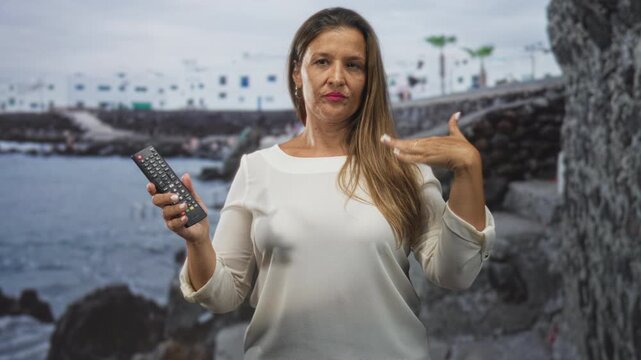 Woman middle aged hispanic holding remote control and touching long hair with right hand in studio seascape backdrop near stone pier; indifference leisure.