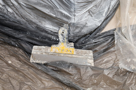 An old dirty construction spatula with remnants of dry putty and plaster lies on a protective plastic film. A wide metal tool with a yellow handle was used during apartment renovation or interior fini