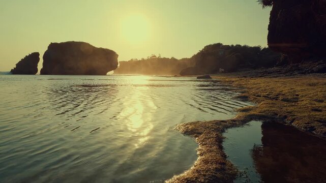 A serene low-angle shot of ocean waves gently washing onto a moss-covered rocky shore at golden hour with distant karst islands silhouetted against the warm glowing sun.