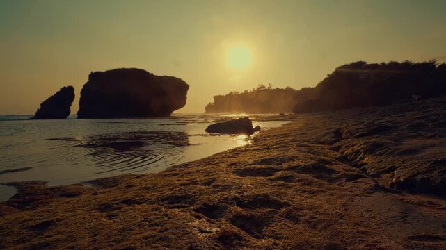 Serene coastal scenery featuring dramatic rock formations silhouetted against a vibrant golden sunset, captured with tranquil waves gently lapping at the wet, textured shoreline.