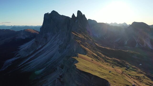 Breathtaking high-angle drone shot of the dramatic Seceda peak and sloped alpine meadows in Val Gardena, Italy. Features majestic jagged mountain ridges illuminated by warm golden hour sunset light.