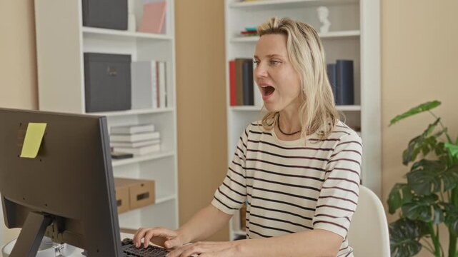 Woman at computer smiling and raising clenched fists in a victory pose while seated at desk with keyboard and monitor in studio; remote work success joy.