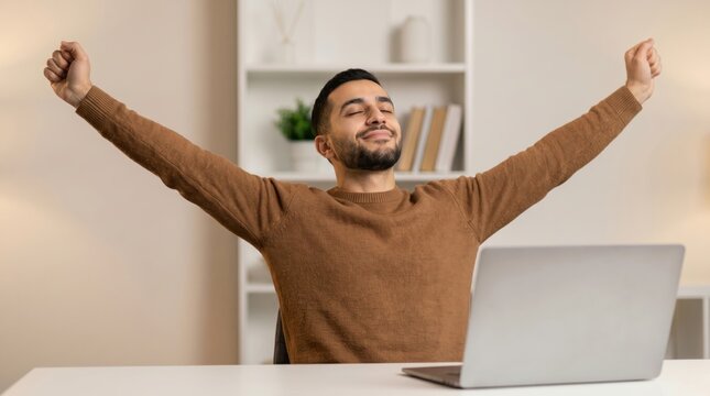 Happy young man stretching arms after finishing work on laptop at home office.