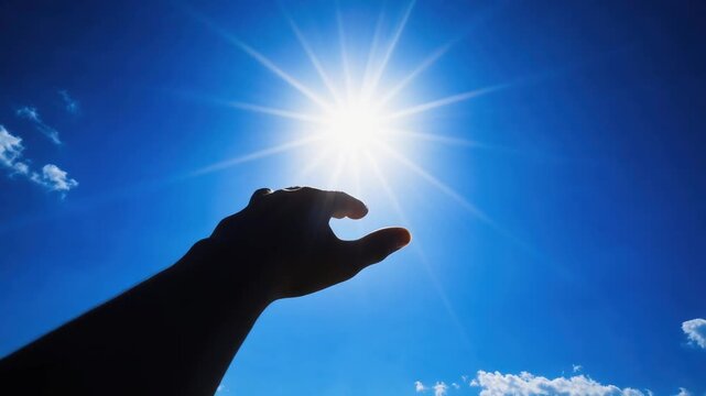 Silhouette of hand reaching towards bright sun in clear blue sky with few clouds on a sunny day