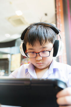 Asian schoolboy wearing glasses and headphones looking at tablet screen. Concept of digital e-learning, online gaming, childhood technology, modern education, and indoor entertainment.