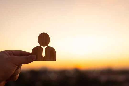 Hand holding a wooden businessman figure against a bright sunset sky. Concept of human resources, successful recruitment, leadership, career opportunity, and business future growth.