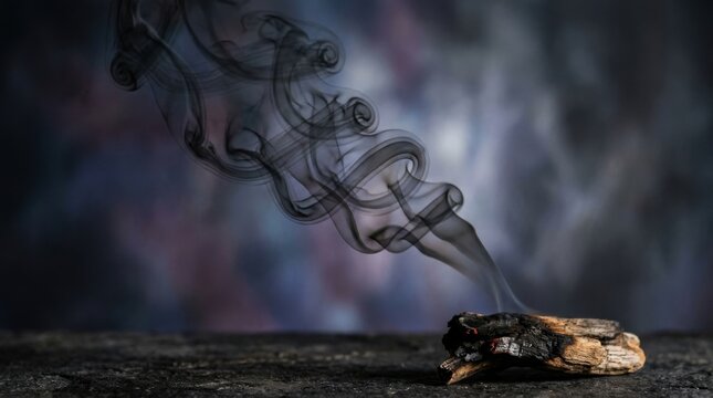 Dark swirling smoke patterns rising from a smoldering piece of charred wood on a stone surface with glowing embers against a moody textured dark background with bokeh.
