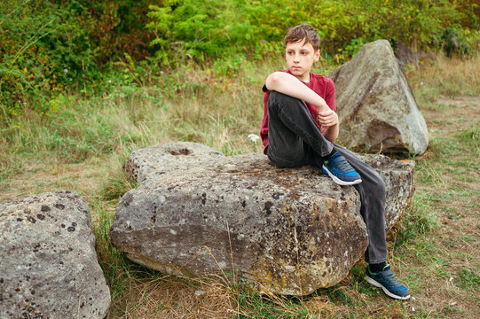 Thoughtful teenager boy sitting on large rock in countryside landscape