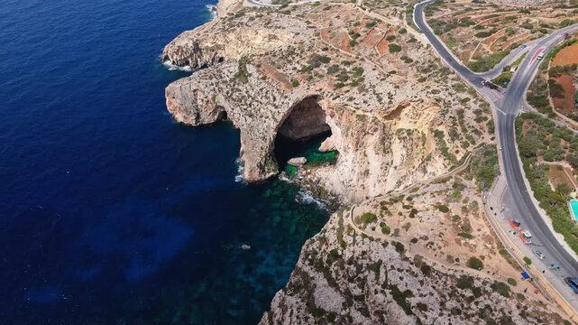 Malta&rsquo;s Blue Grotto, a majestic natural limestone arch and sea cave complex on the southern coast. Sweeping aerials show deep azure waters contrasting with rugged, sun-drenched cliffs.