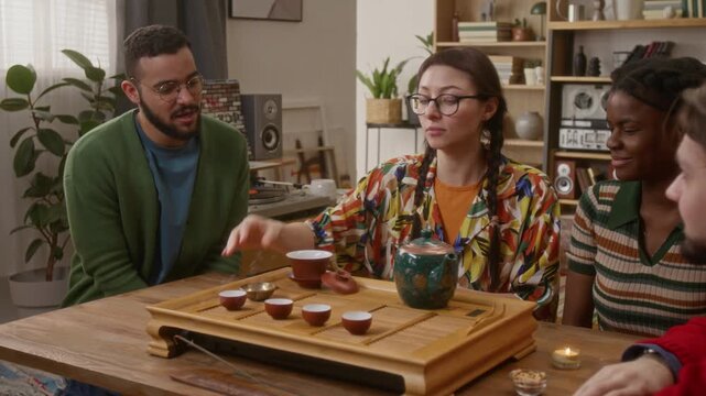 Young woman in tropical shirt intently pouring water from turquoise teapot into teacup, while three friends watching ritual with warm smiles