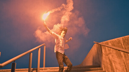 A skateboarder in adolescence ignites a flare, casting a vivid glow as he stands on a stair railing at dusk, embodying rebellious energy and youthful exuberance. © wkproduction