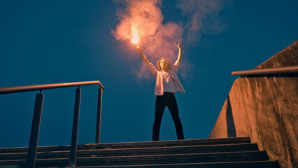 A young skateboarder stands triumphantly on a stair set, holding a flare in one hand, surrounded by a vibrant cloud of smoke against a deep blue sky. © wkproduction