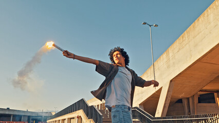 A young skateboarder stands on a rooftop holding a flaming torch, embodying the carefree spirit of youth culture. The late afternoon sun highlights his casual attire and the urban backdrop. © wkproduction