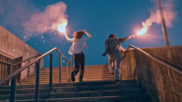 In the serene dusk, two adolescents, one caucasian with curly hair and another of indeterminate ethnicity, energetically descend a staircase, each holding a flaming flare.