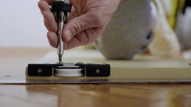Closeup hands securing roller with screwdriver, white technician aligns bracket and swaps bits on parquet floor, focused renovation scene, precise tool handling, modern DIY energy