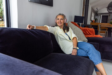 Fototapeta premium Senior woman relaxing sideways on dark velvet sectional at home, wearing pink headphones, smiling