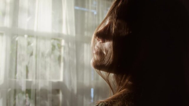 Over shoulder close up shot of summer breeze blowing onto young woman standing by window at home with wind ruffling long hair, copy space