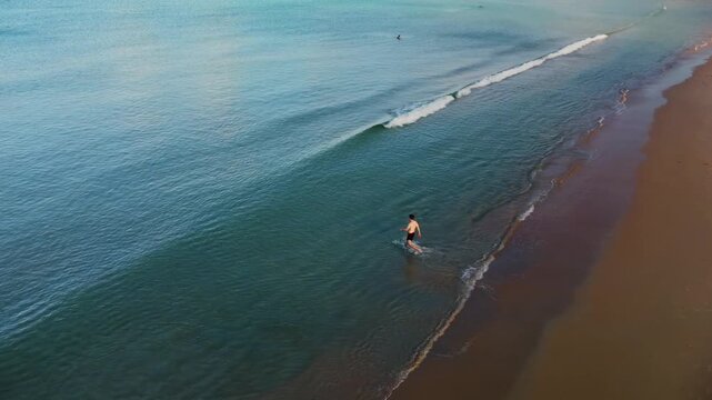 Caucasian man is walking into azure waters among calm turquoise waves on sandy beach and starting to swim. Aerial view of tranquil coast with holiday maker amidst sparkling blue ocean. Journey theme