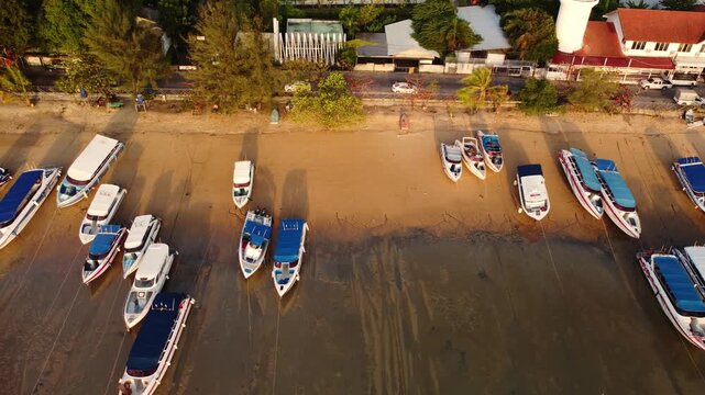 Overhead boats resting on wet sand at low tide with colorful hulls lined on exposed beach. Fishermen ships near village rooftops and palm fringe in background with long shadows. Thailand shoreline 