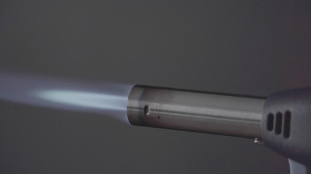 Close-up of a gas torch nozzle. Bright blue flame emanates from the tip. Flame is steady and intense. Neutral gray background keeps focus. Ideal for culinary or industrial heating demonstrations
