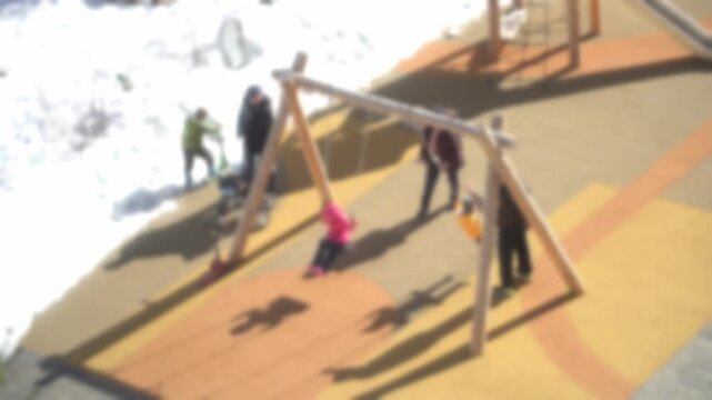 Snowy playground has a wooden swing set. Children play joyfully on the swings. Pink-clad girl swings high. Another child in yellow stands nearby. Adults watch closely. Kids smile broadly