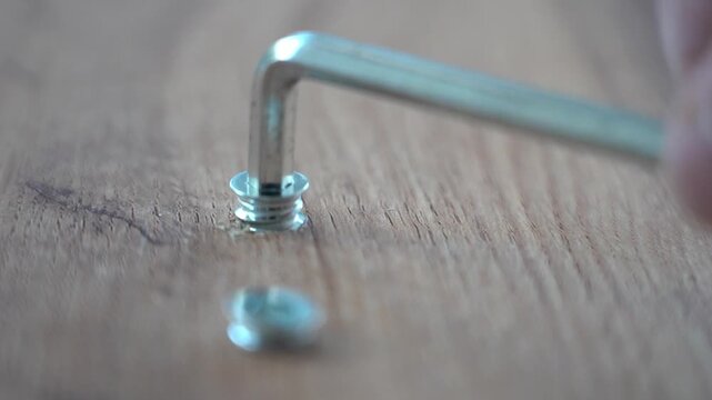 Close-up of a hand using a hex key on wood. Two metal fasteners rest on the grainy surface. The hand moves with focused precision. Ideal for DIY or repair projects