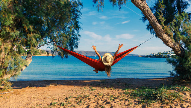 happy woman in straw hat relaxing on a red hammock by the beach, summer vacation and freedom concept