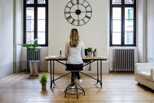Rear view of woman sitting at desk at home under large wall clock