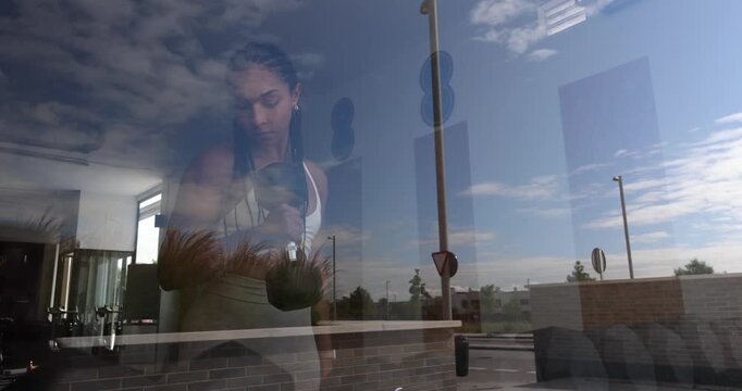 Determined african american woman performing bicep curls with a dumbbell, seen through the gym window with the sky and clouds reflected on the glass, showcasing strength and commitment