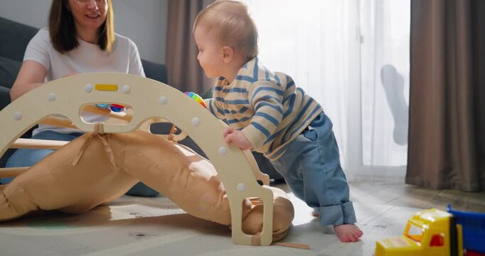 A toddler wearing a striped sweater and blue pants is playing and experimenting with a wooden climbing arch toy in a sunlit living room while a woman closely supervises and encourages the child.