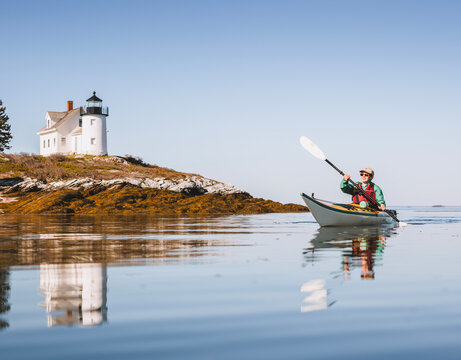 Woman sea kayaks on calm water near lighthouse in Maine.