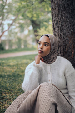 Pensive young muslim woman in hijab relaxing under tree in park