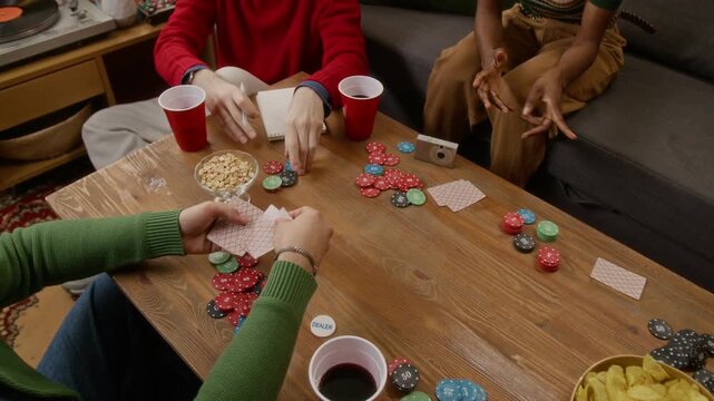 High angle zoom out shot of young adult man distributing cards while playing poker with friends at wooden coffee table with drinks and snacks in cozy living room