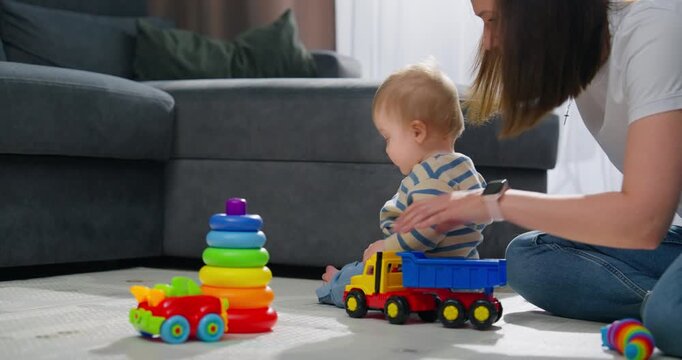 A toddler playing with colorful stacking rings and toy vehicles on a living room floor while being gently supported by an adult during early development and playtime