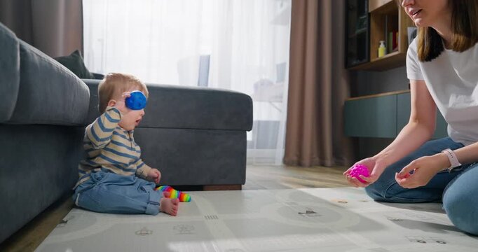 A toddler sitting on the floor engaged in playful interaction with an adult holding colorful toys, demonstrating early childhood play and learning in a cozy living room environment with soft natural