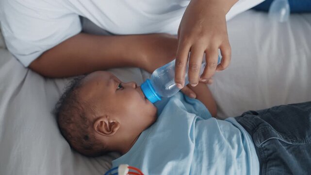 Little child drinking water from bottle, mother helping to infant, mother care. Feeding and taking medicine, curing infants with modern medicine, african american baby boy lying in bed and go to sleep